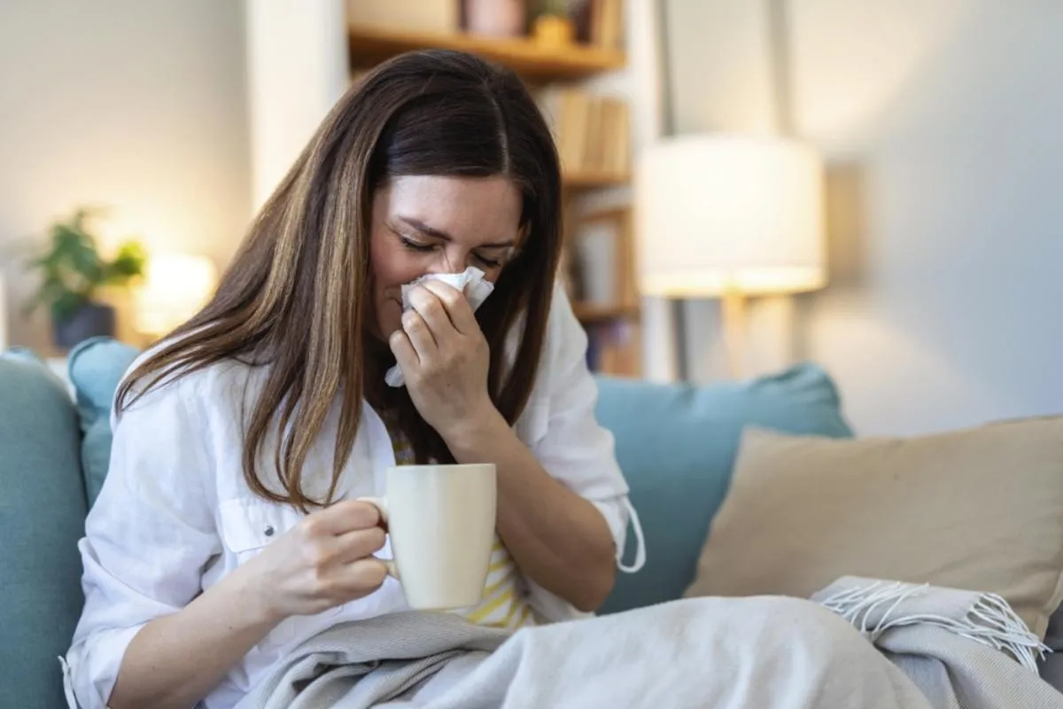 A megfázás enyhe tünetekkel jár: egy beteg nő ül a kanapén, fújja az orrát és forró italt tart a kezében