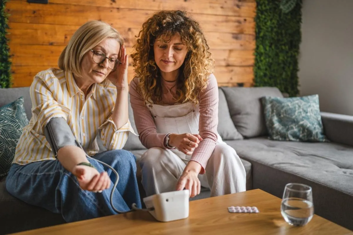 Magas vérnyomás: egy idősebb nőnek méri egy fiatalabb a  vérnyomását az otthonukban egy kanapén
