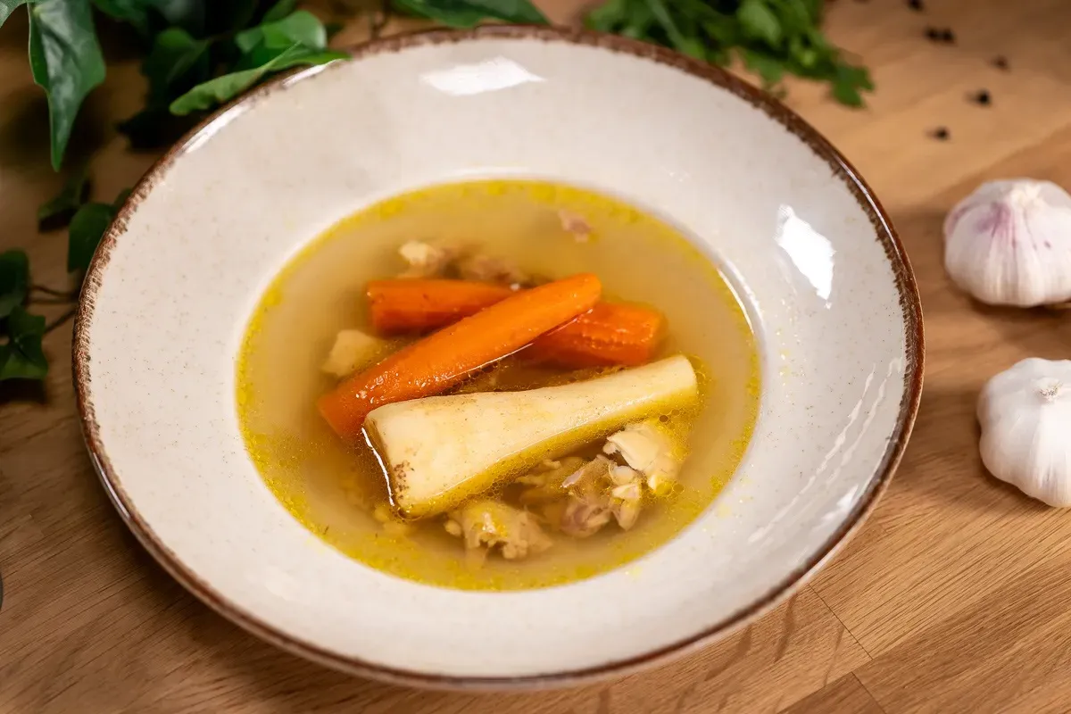 Egy fehér tányérban aranyló húsleves petrezselyemgyökérrel, répával és hússal