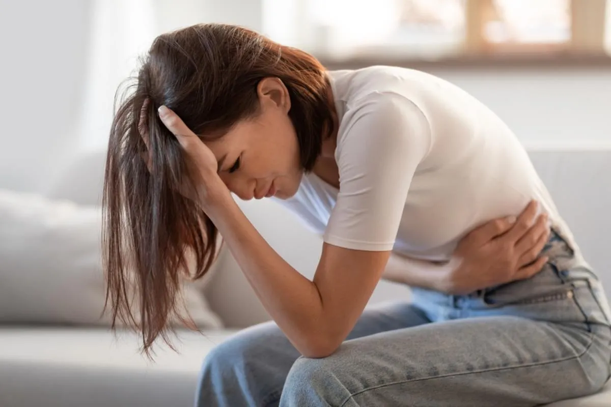 Endometriózis tüneteit mutató nő ül az ágyon, hasát fájlalja.